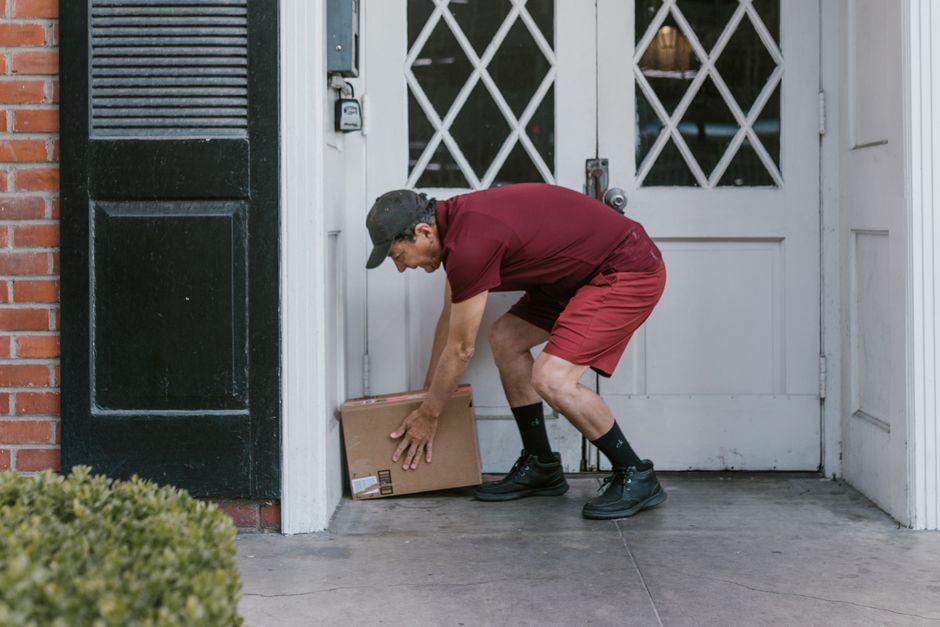 A delivery person from floristdowne.co.uk is shown placing a cardboard box on the ground in front of a white door with diagonal-paneled glass. The person is wearing a dark grey cap, a maroon t-shirt, 