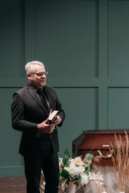 The image features a man with light-colored hair and glasses, dressed in a black suit and shirt, standing indoors against a muted teal wall with rectangular paneling. He is holding an open book or boo