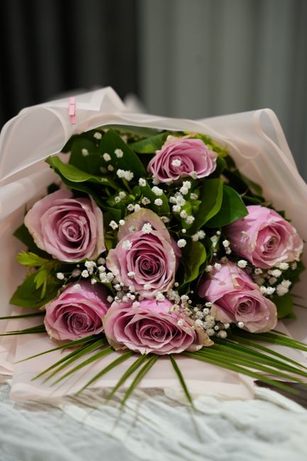 A bouquet of fresh, pastel pink roses with velvety petals and tightly packed blooms, complemented by small white baby's breath flowers and lush green leaves. The arrangement is wrapped in elegant whit