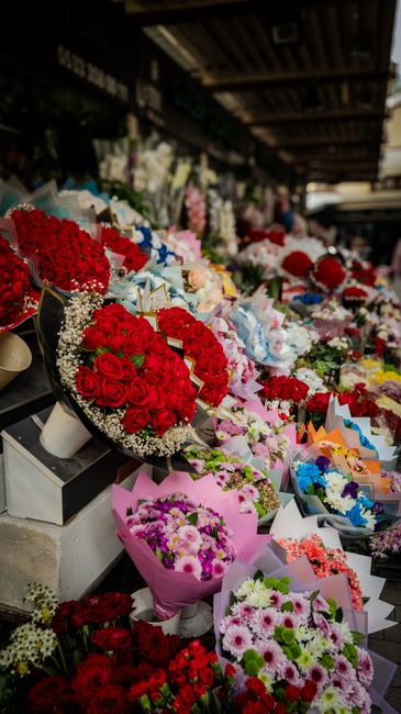 An outdoor flower stall displaying a vibrant array of fresh floral arrangements and bouquets arranged on tiered shelves under a wooden canopy. Prominent red roses are gathered into round bouquets wrap