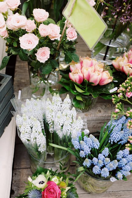 A display of fresh flower bouquets arranged on a rustic wooden table in a florist shop. The arrangement includes a cluster of pale pink roses with lush, dark green leaves in a tall, clear glass vase. 