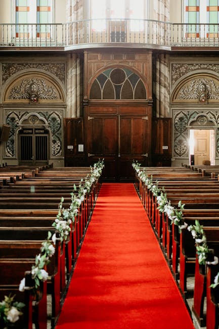 A view of an elegant church interior prepared for a wedding ceremony, featuring a central aisle with a vibrant red carpet leading to a large wooden double door at the altar. The aisle is flanked by da