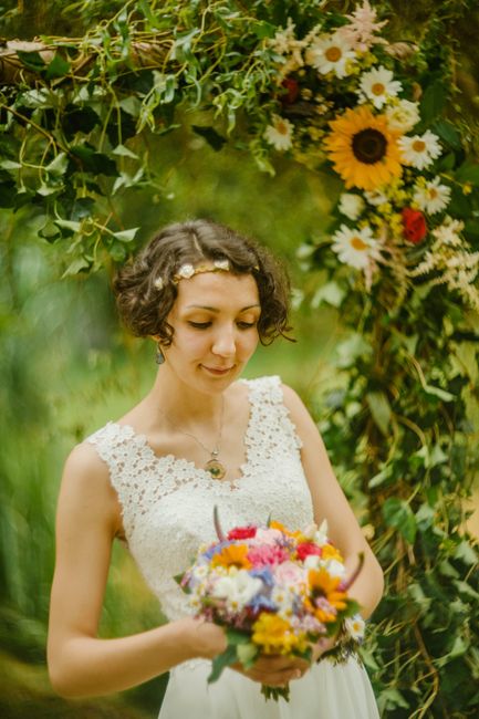 A young woman with short, curly brown hair and a delicate flower headband is holding a vibrant bouquet of fresh flowers, including yellow, pink, purple, and white blooms, during an outdoor wedding cer