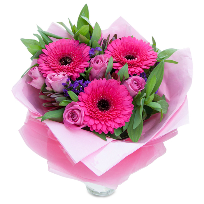 A composition that marries playful color with gentle grace: three bold gerbera daisies in vivid fuchsia sit at the center, their many petals unfurling like joyful suns around deep dark centres. Nestled close by, roses in a dusty lavender-pink tuck their velvety petals inward, creating a tender counterpoint to the gerberas' exuberance. Small clusters of purple statice or similar filler flowers pepper the spaces between, their papery texture catching light and adding a whisper of wildflower charm. The foliage is generous and verdant - broad salal leaves and slender eucalyptus providing structure, a cool green canvas that makes the pinks sing and gives the bouquet a fresh, just-gathered-from-the-garden feel.

The arrangement is wrapped for giving, starting with a soft, matte pale-pink tissue that cradles the stems, and finished with a sheer, slightly crinkled fuchsia-pink cellophane that shimmers faintly and ties the colour story together. The top rim of a clear glass vase is visible below, suggesting this piece is ready to be displayed at once. In the gentle daylight you might find in Downe - whether after a walk around the village green or a visit to the nearby lanes toward Cudham - the roses' mild perfume mingles with the green scent of eucalyptus to produce an understated, pleasing aroma. This bouquet reads as intimate and celebratory, perfect for marking birthdays, Mothering Sunday, anniversaries, or simply to brighten a neighbour's day with floral warmth.